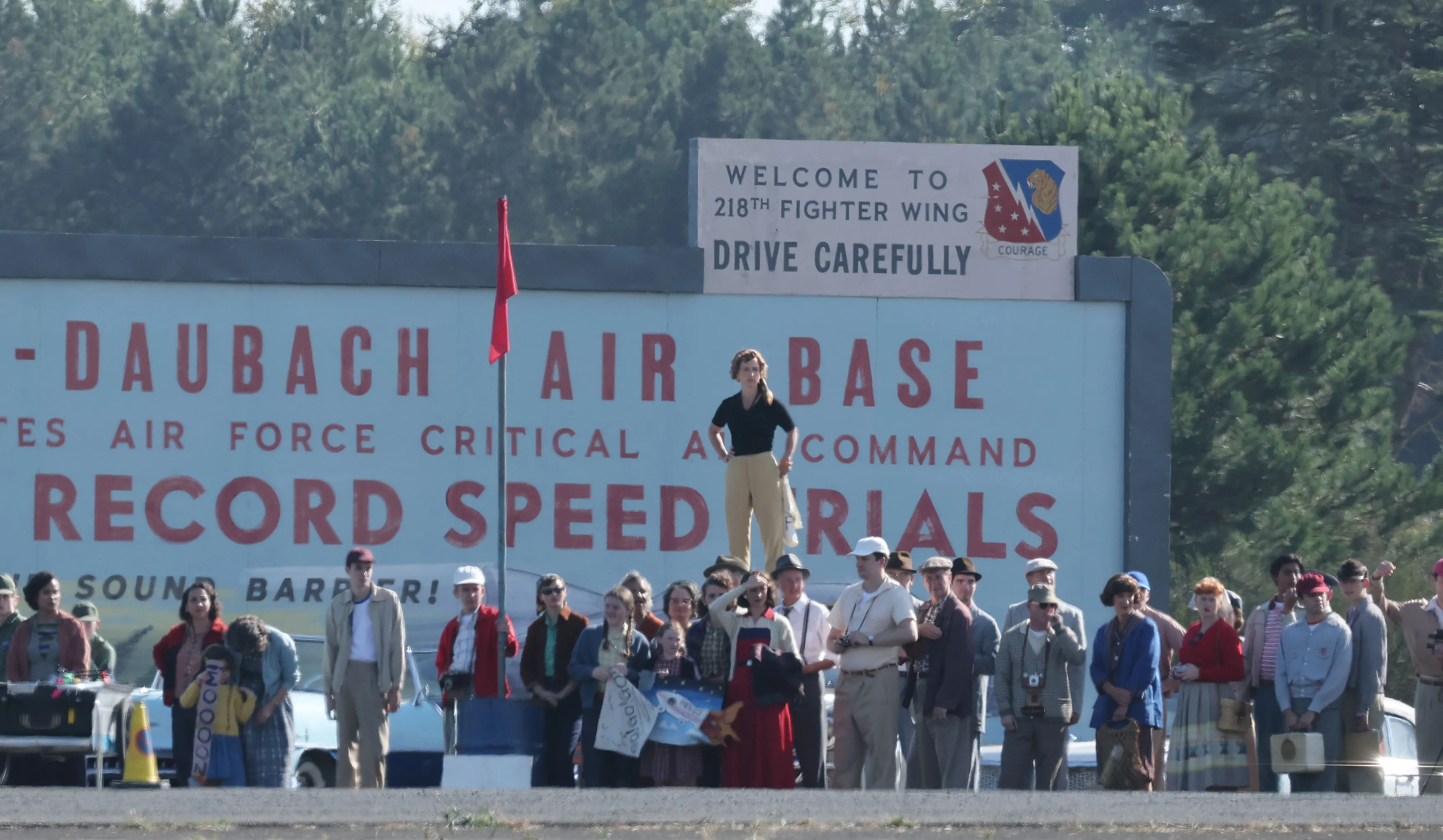 A crowd of people in 1950s attire stand before a sign for 