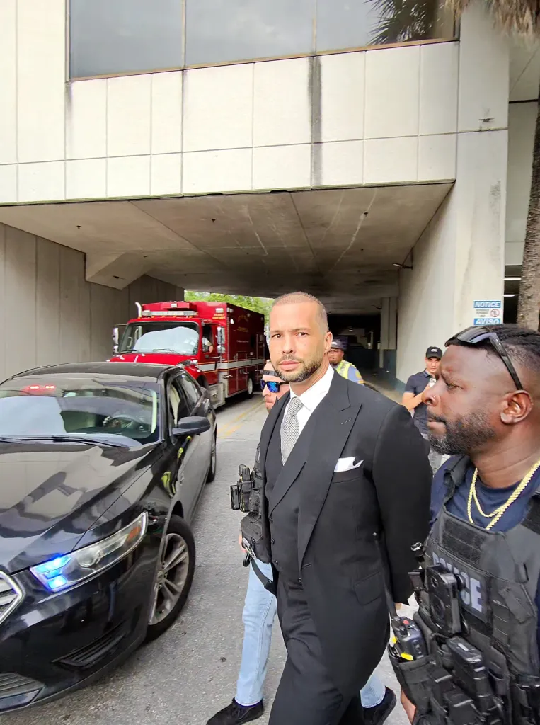 Tristan Tate in a black suit being escorted by an officer in a police vest with a police car and fire truck in the background.