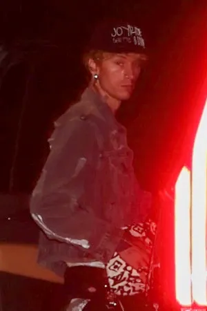 Machine Gun Kelly wearing a denim jacket and a black cap with text on it, looking to his left, illuminated by a red light.