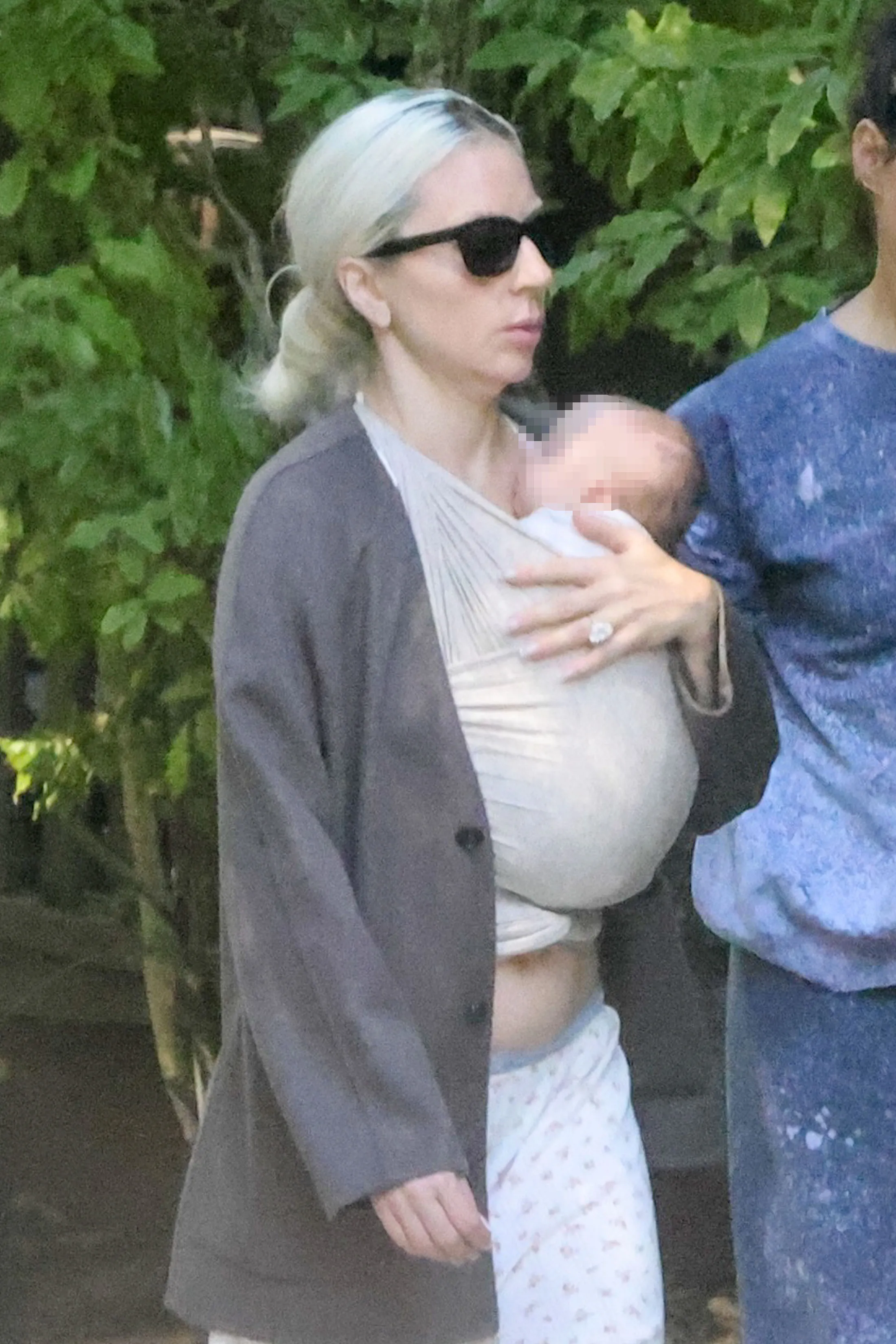 Lady Gaga wearing sunglasses and carrying a baby in a wrap, with a large diamond ring on her left hand.