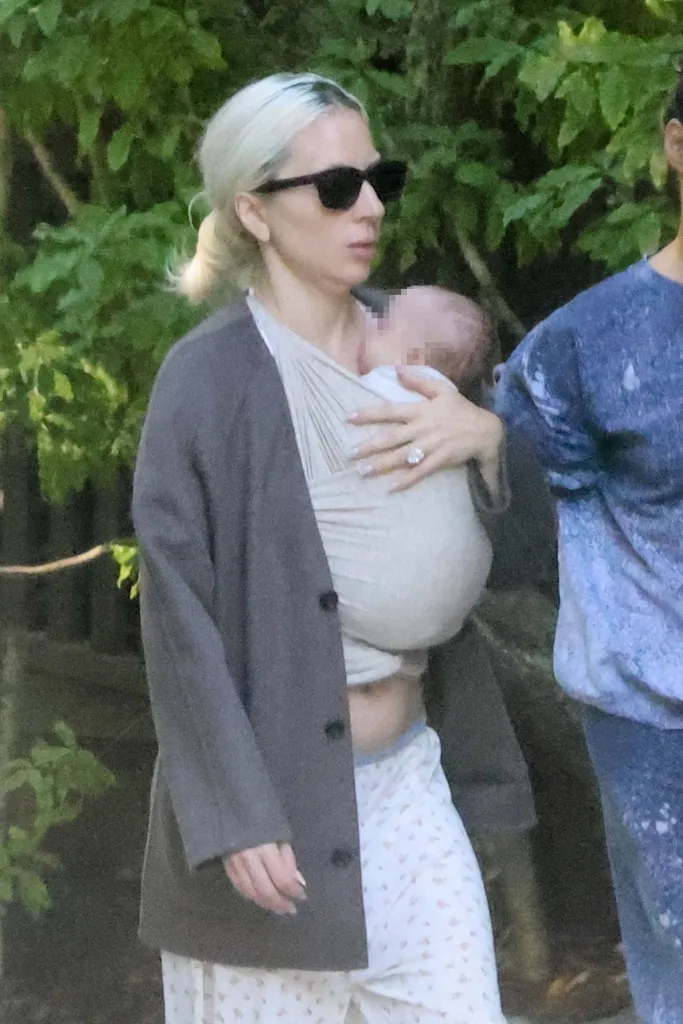 Lady Gaga walking outdoors while carrying a baby in a wrap, accompanied by Michael Polansky.