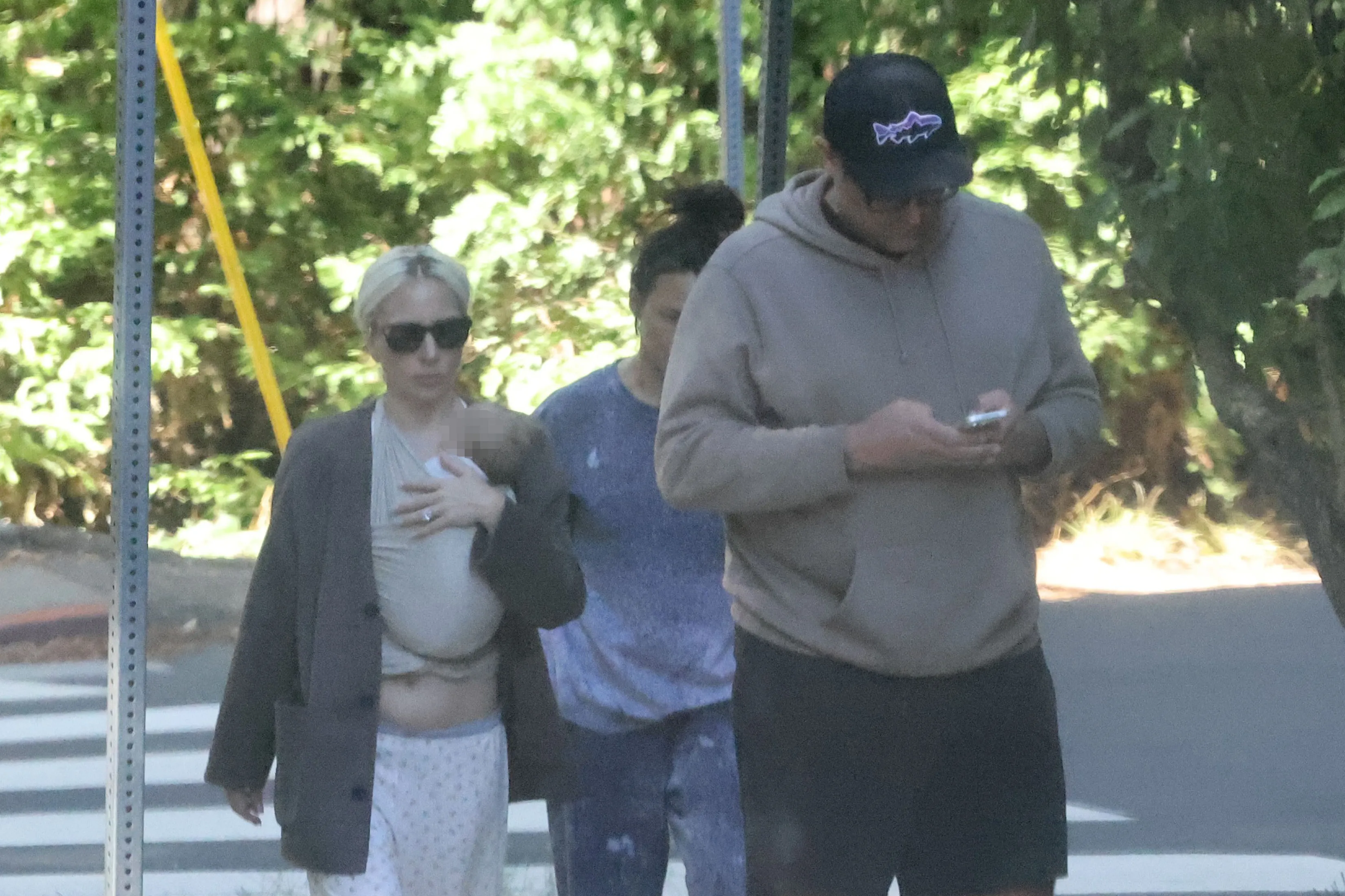 Lady Gaga in sunglasses, wearing a baby carrier with a baby, walking next to Michael Polansky who is looking at his phone.