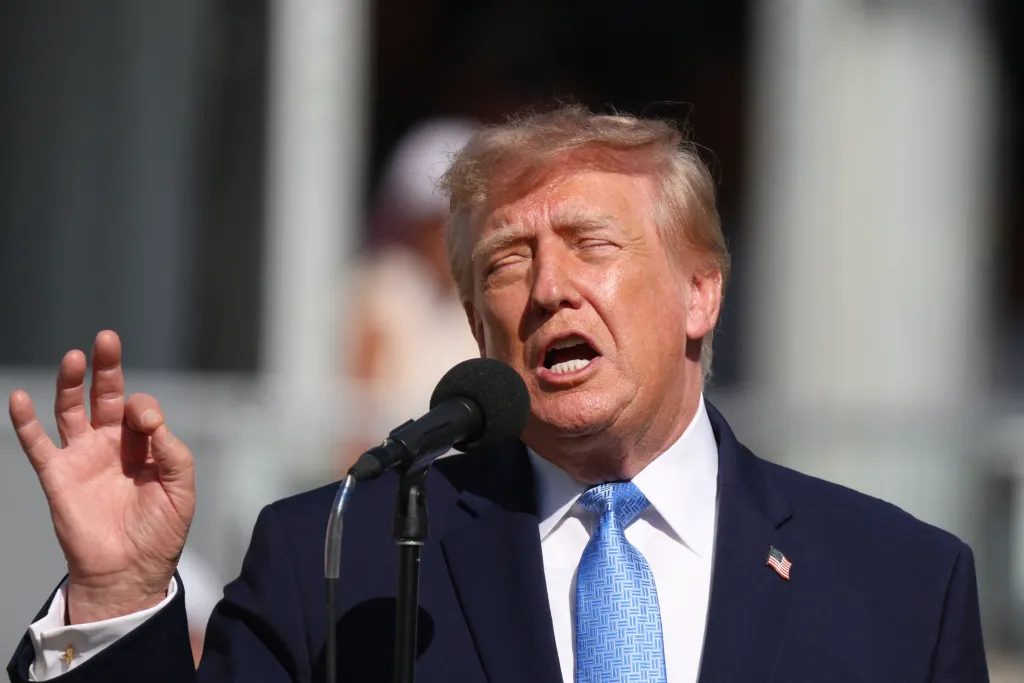 President Donald Trump speaking into a microphone with his right hand raised, addressing the gallery at the Presidents Cup.