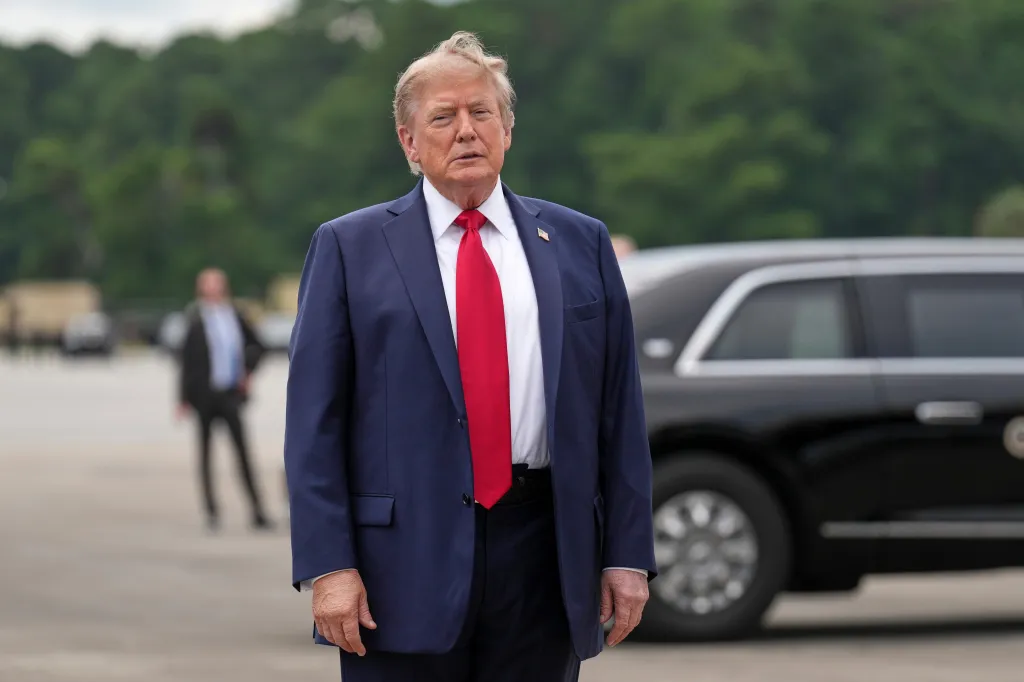 President Donald Trump stands on an airfield in front of a dark car.