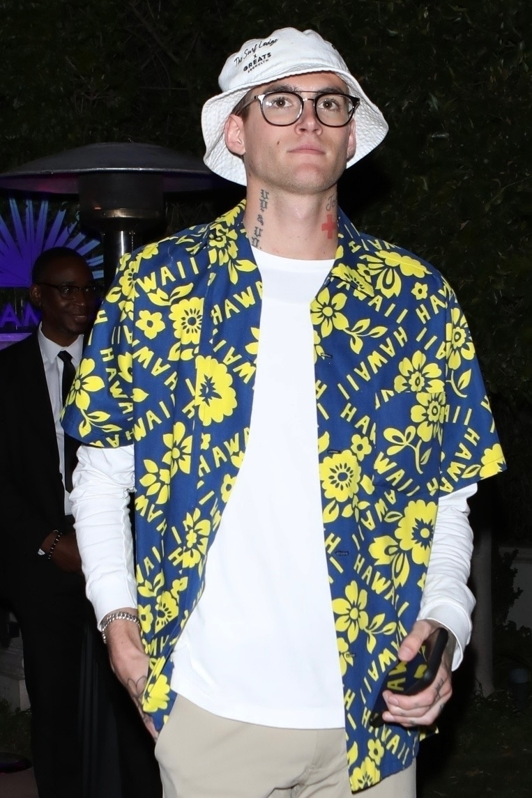 Presley Gerber wearing a blue and yellow Hawaiian shirt, white long-sleeved shirt, and a white bucket hat.
