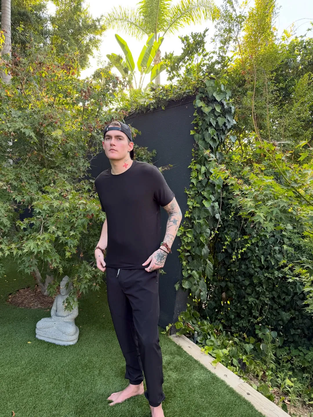 Presley Gerber stands barefoot on grass in a black t-shirt and pants, surrounded by lush greenery.