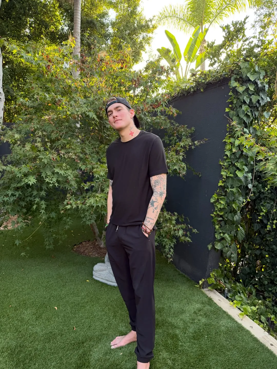 Presley Gerber standing in a garden wearing a black t-shirt, black pants, and a black baseball cap.