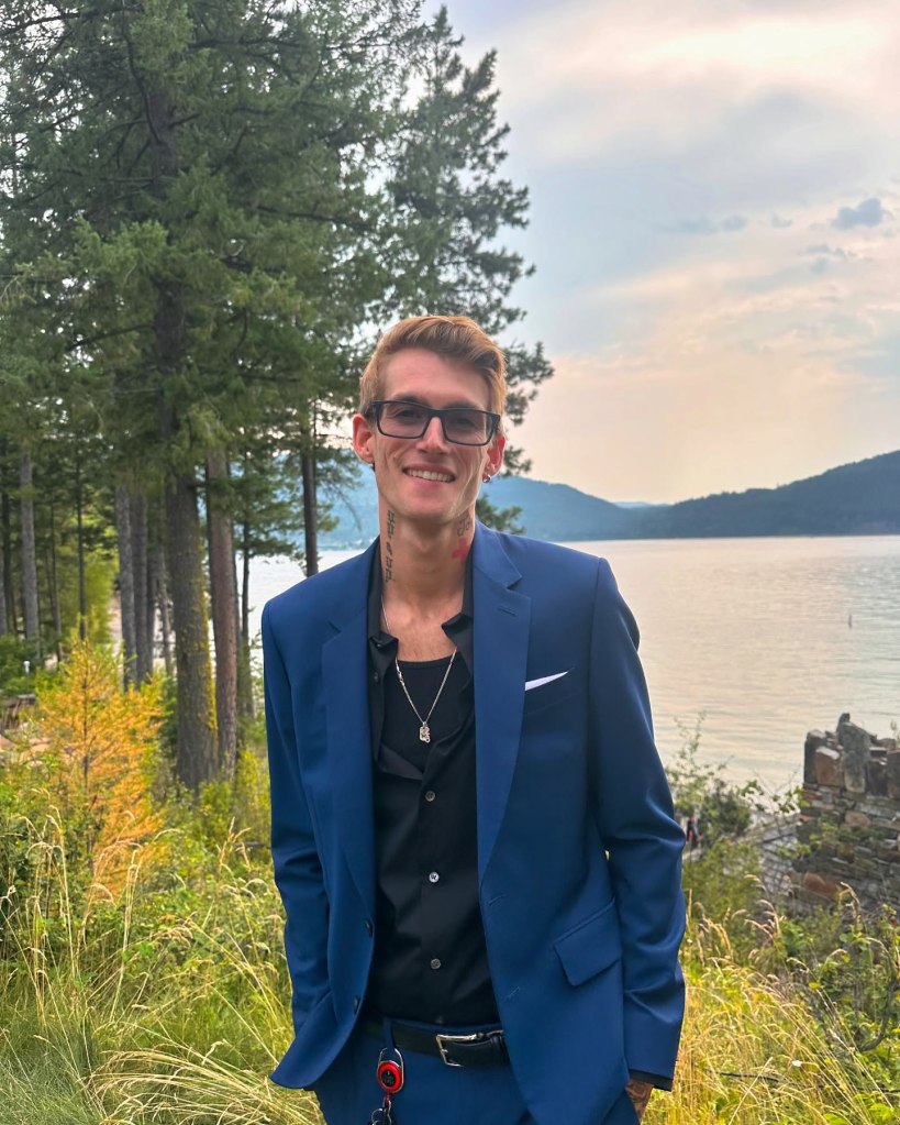 Presley Gerber smiling at the camera, wearing a blue suit and black shirt, against a backdrop of a lake, mountains, and trees.