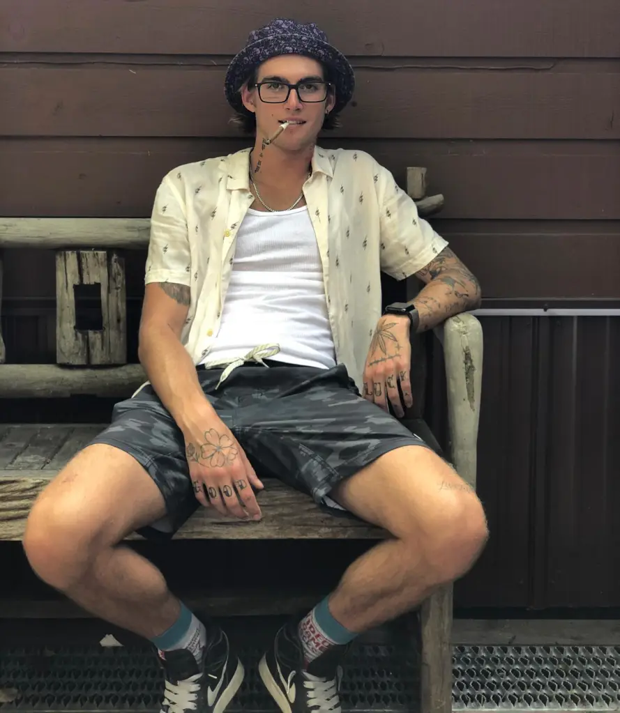 Presley Gerber, model and son of Cindy Crawford, sitting on a wooden bench with a cigarette in his mouth.