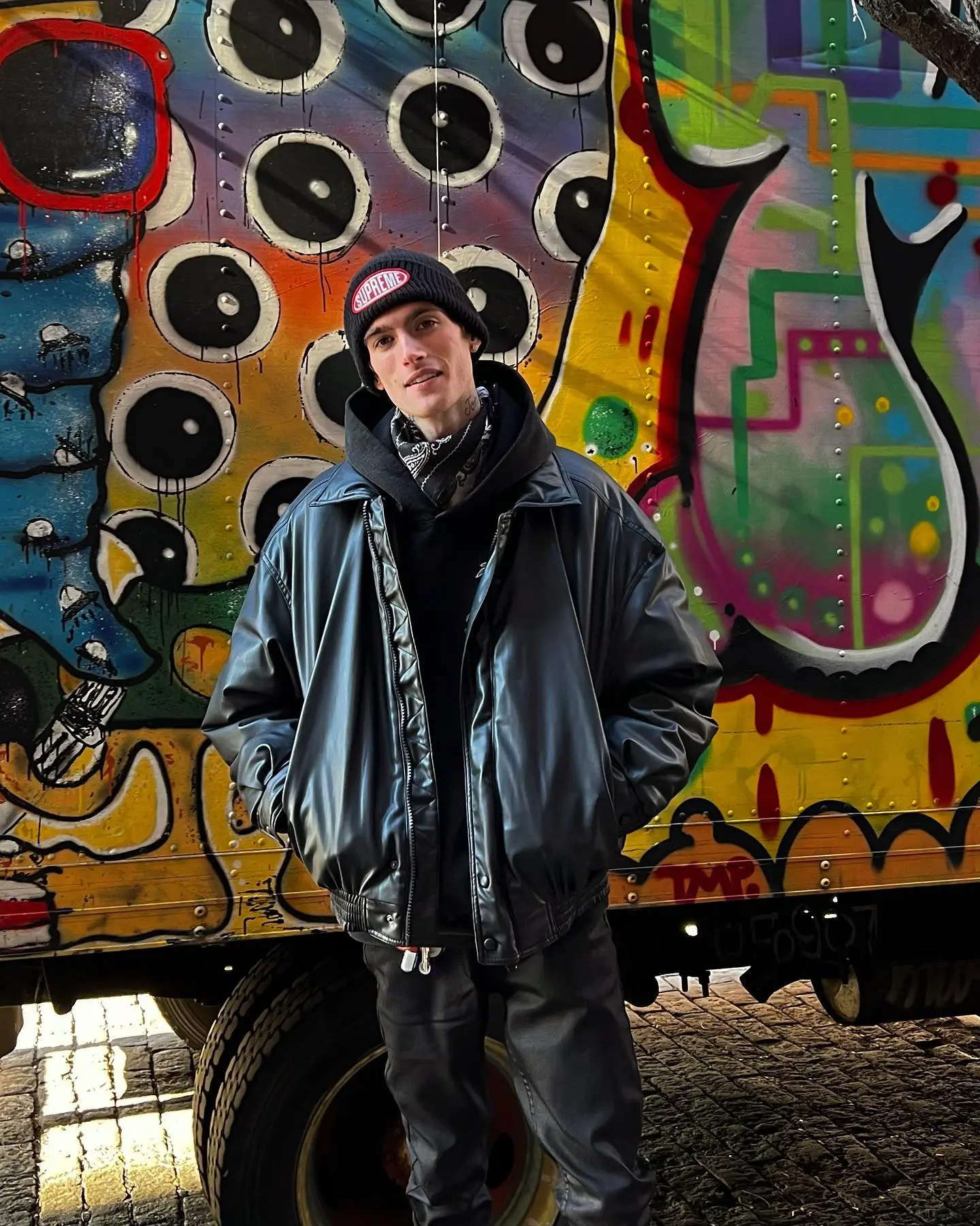 Presley Gerber in a black outfit and Supreme beanie, standing in front of a colorful, graffiti-covered truck.