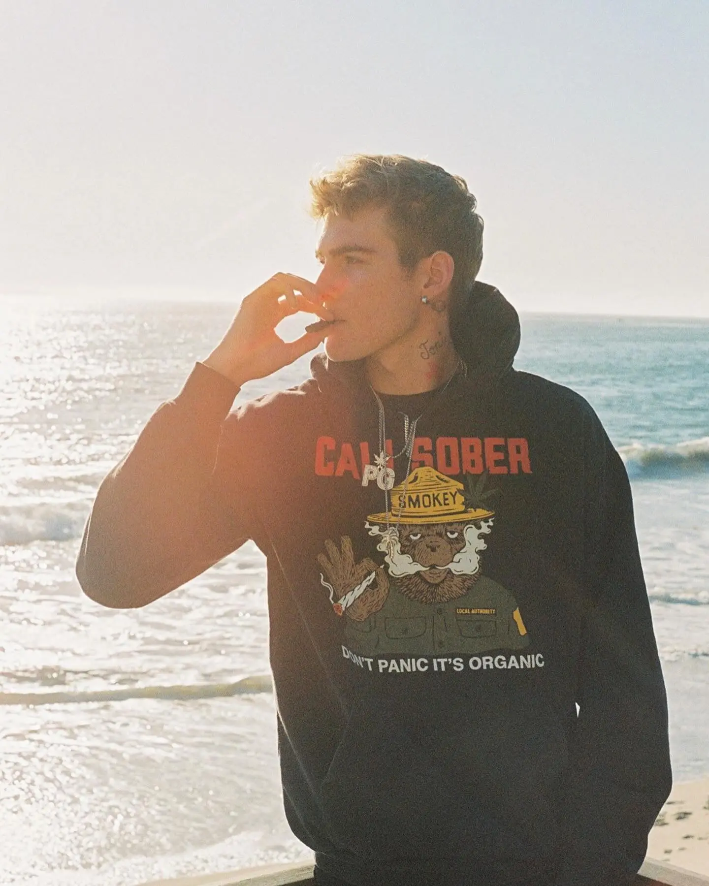 Presley Gerber wearing a black hoodie with a Smokey Bear graphic and smoking by the ocean.