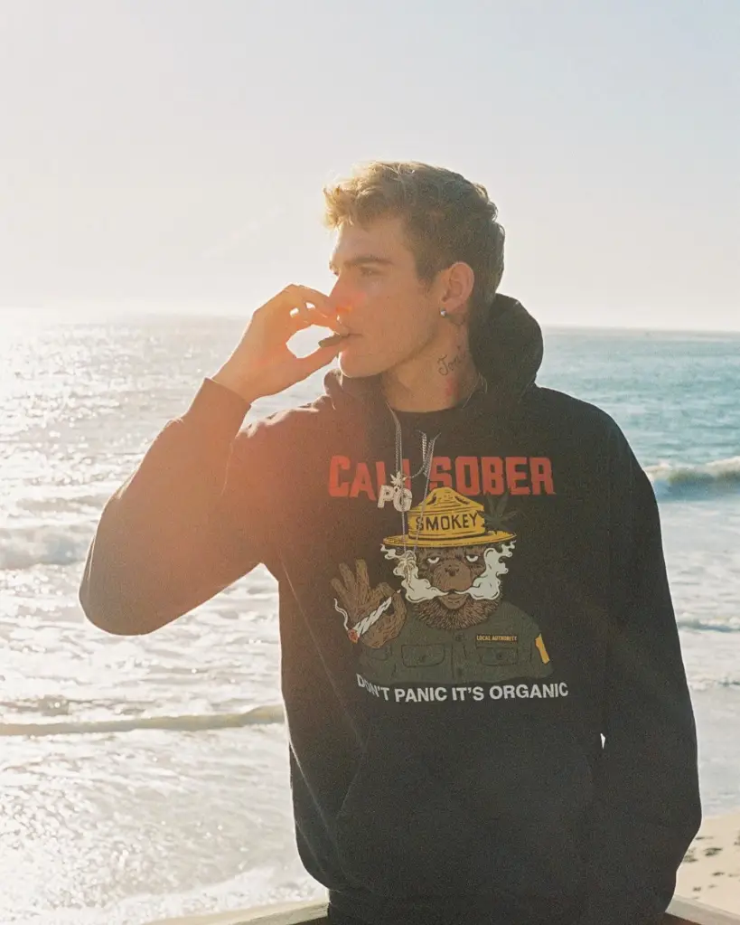 Presley Gerber, model son of Cindy Crawford and Rande Gerber, in a black hoodie smoking a joint, with the ocean in the background.