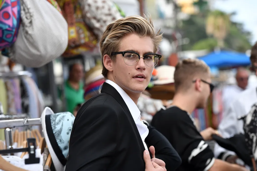 Presley Gerber in a white shirt and black suit jacket, wearing glasses, looking over his shoulder.