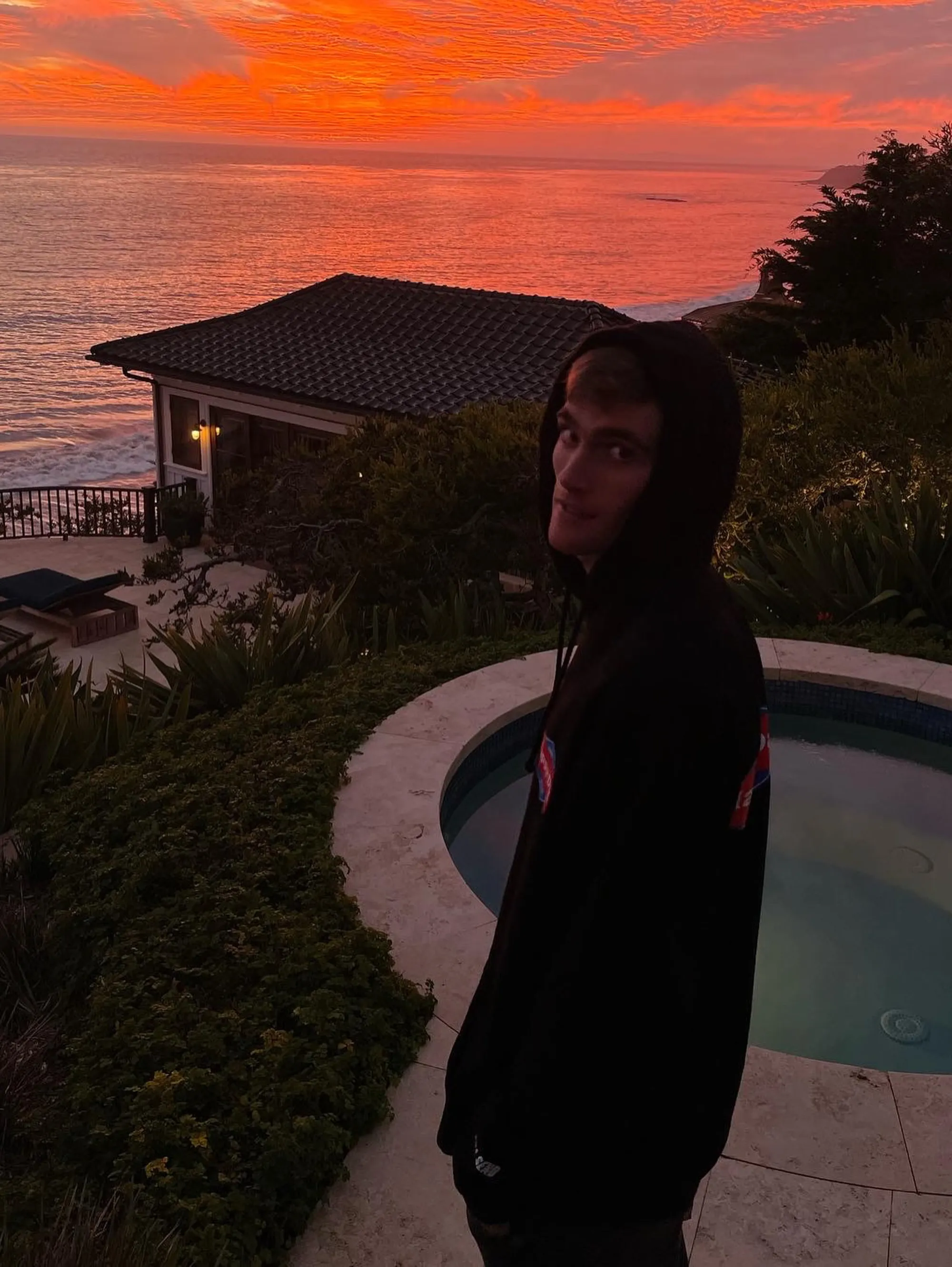 Presley Gerber in a black hoodie by a pool overlooking the ocean at sunset.
