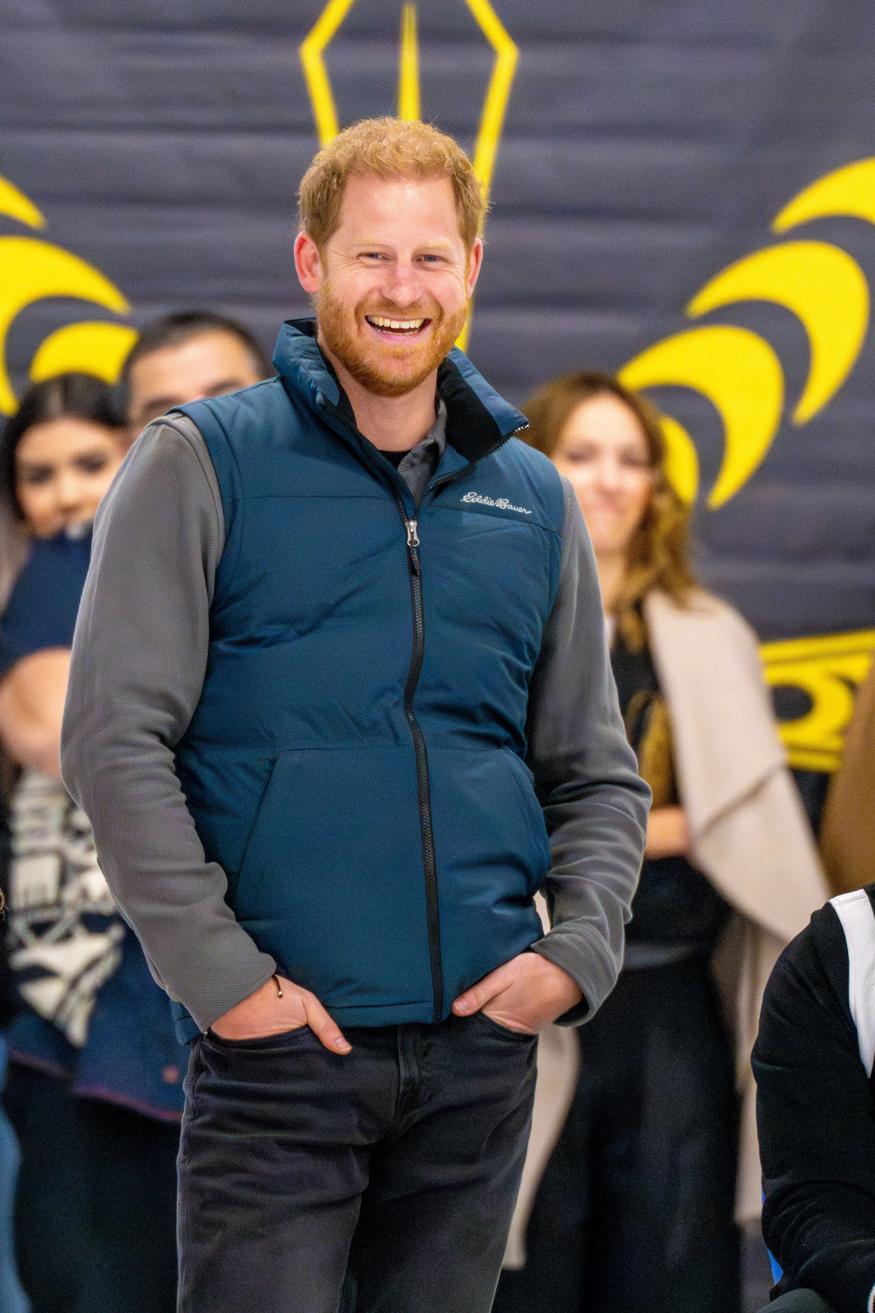 Prince Harry smiling, wearing a blue vest and gray long-sleeved shirt with his hands in his pockets.