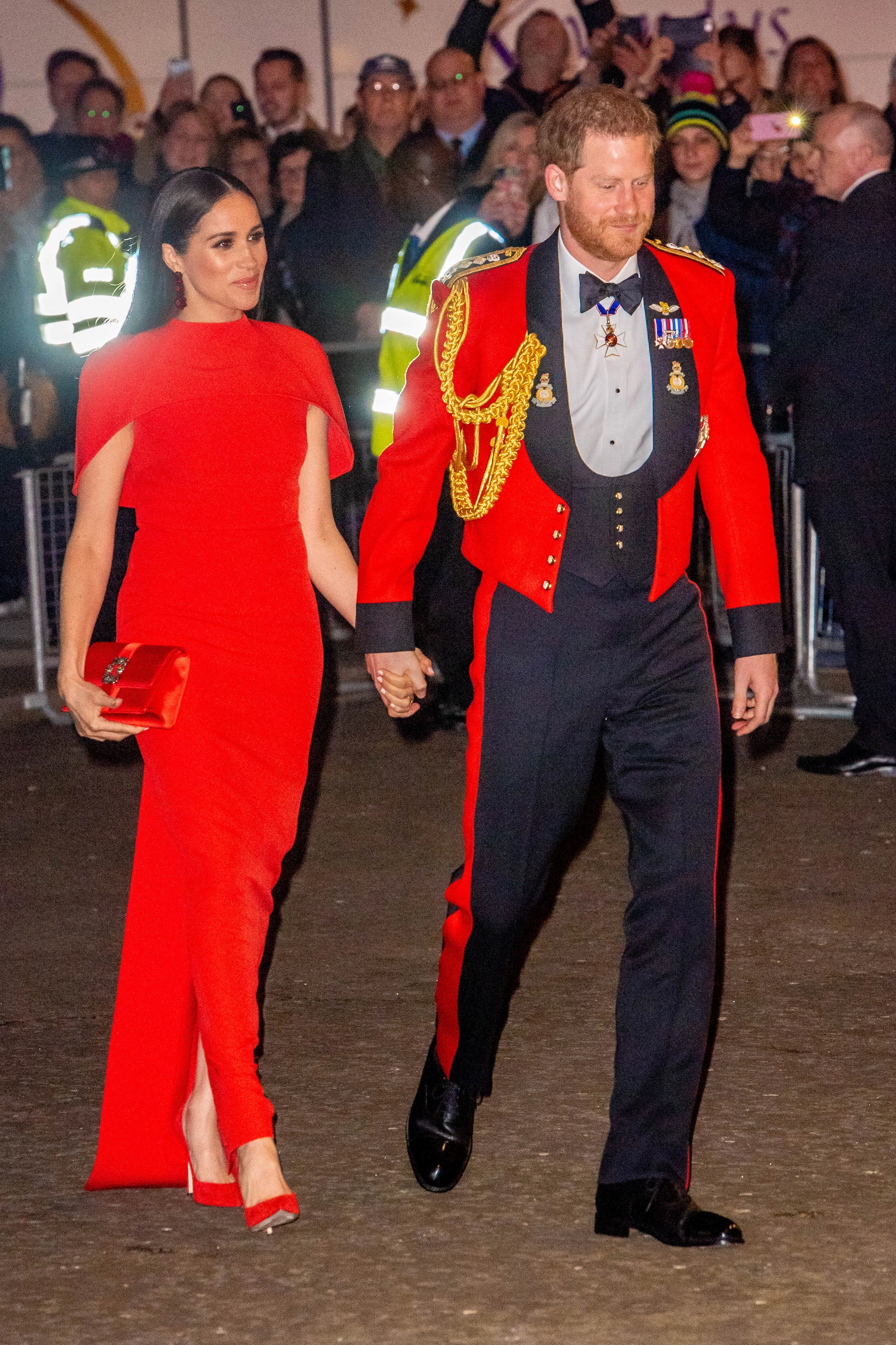 Prince Harry in military uniform and Meghan Markle in a red dress holding hands.