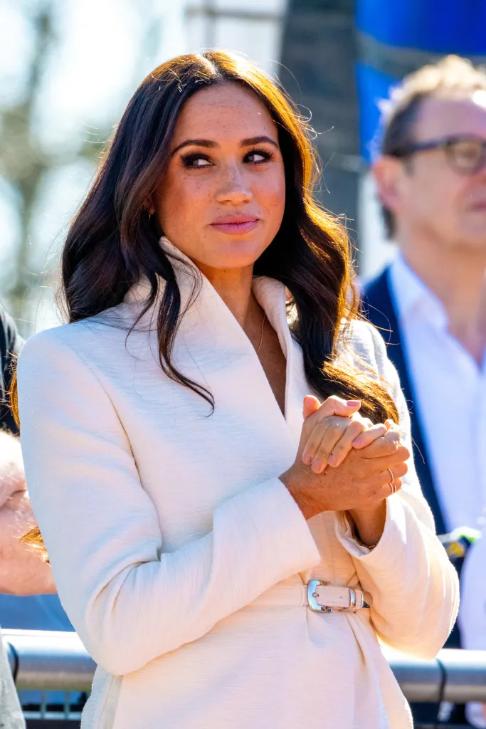 Meghan Markle, Duchess of Sussex, in a white coat, looking to her right with hands clasped.