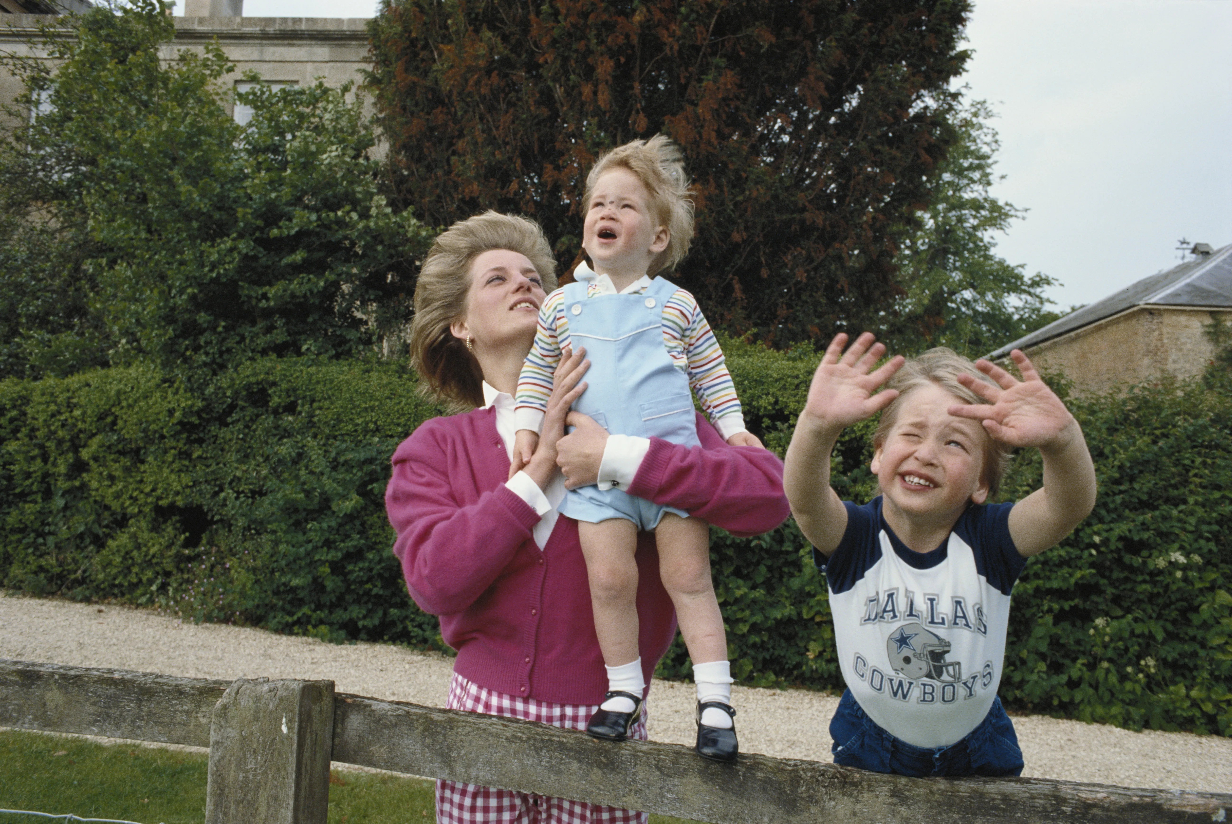 Princess Diana holding Prince Harry while Prince William leans on a wooden fence, smiling.