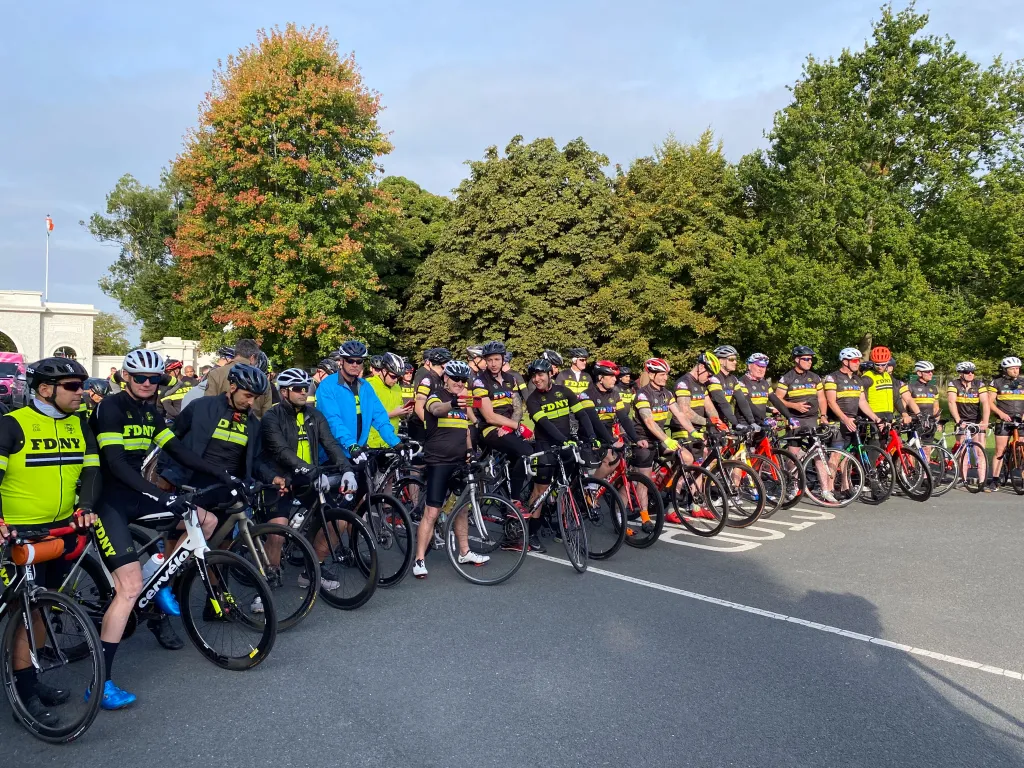 A group of cyclists wearing matching black and yellow FDNY shirts gathered with their bikes on a road.