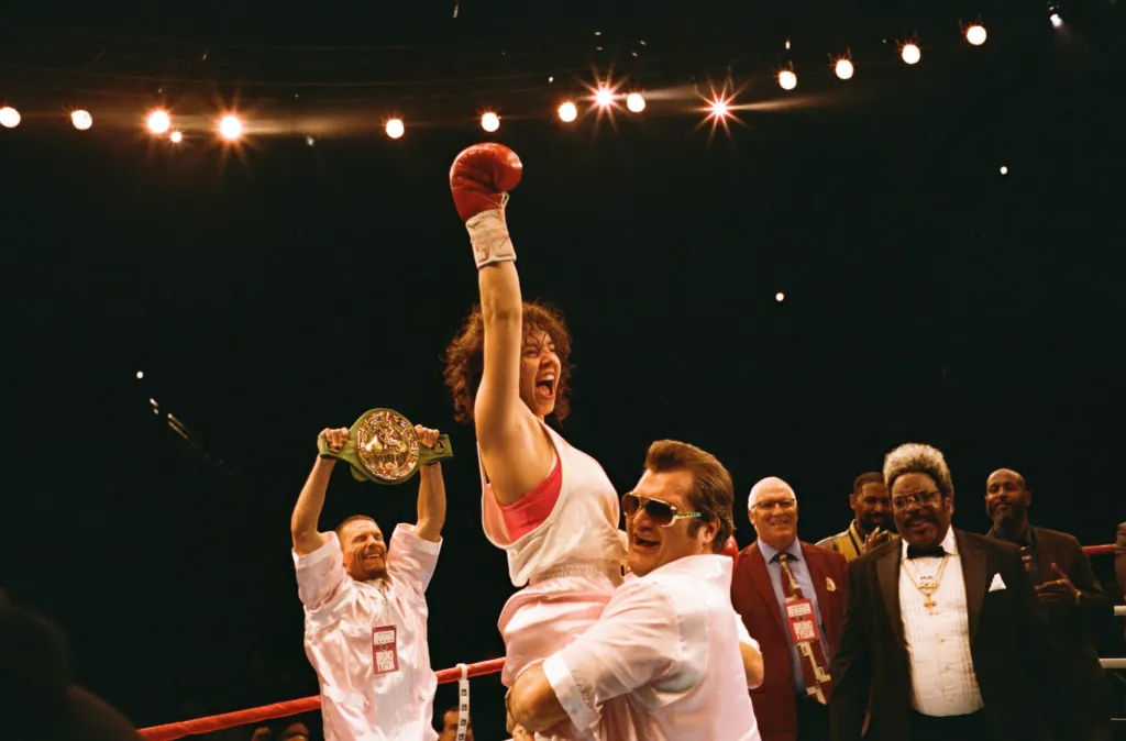 Sydney Sweeney as Christy Martin, in a white tank top and red sports bra, celebrates with a raised glove in a boxing ring, held by another man, as Chad L. Coleman as Don King smiles in the background.