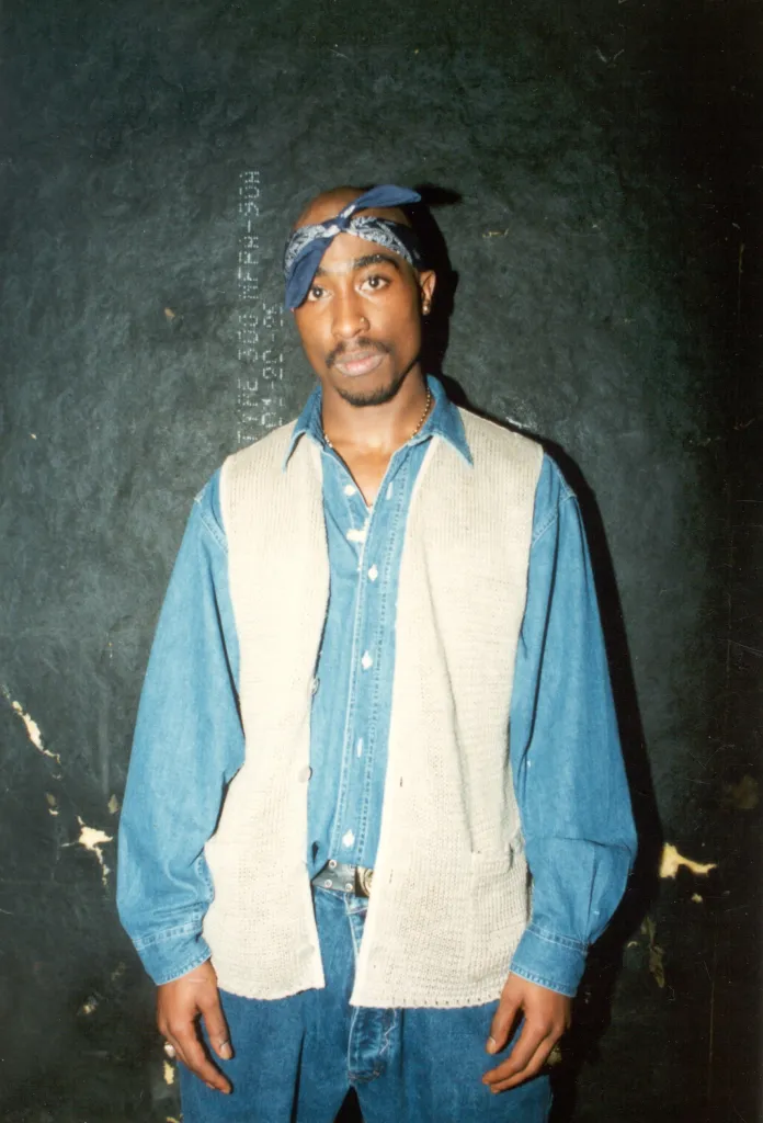 Rapper Tupac Shakur wearing a denim shirt, jeans, and a cream-colored vest, with a blue bandana tied around his head.