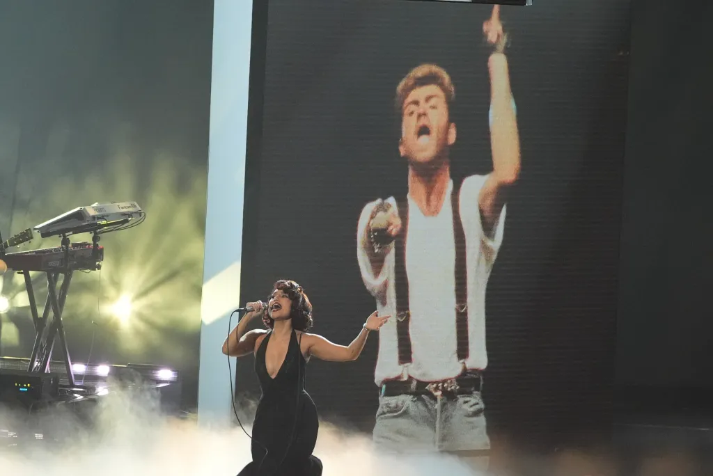 Raye performs on stage in a black dress, singing into a microphone, with a large screen behind her displaying an image of George Michael with his arm raised.