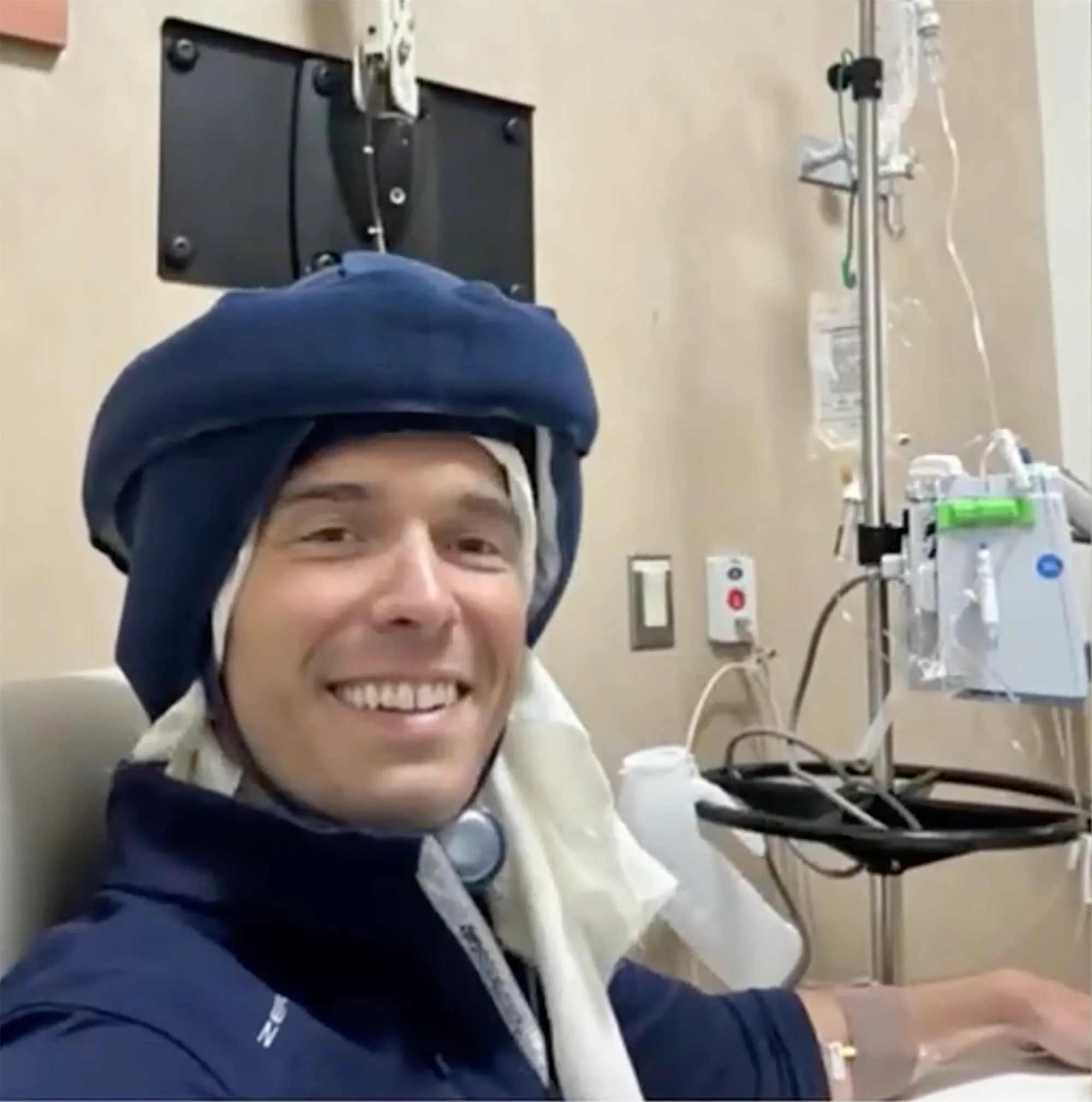 Will Reeve smiling while wearing a cold-cap therapy helmet in a hospital room with medical equipment in the background.