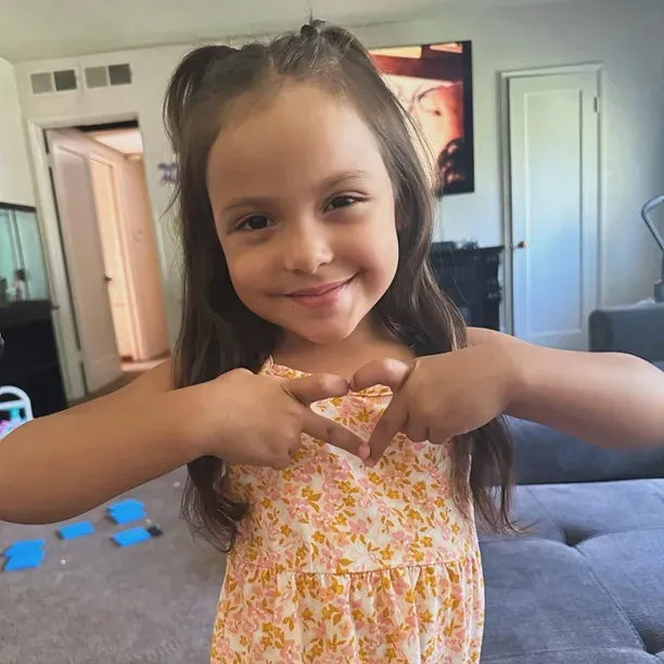 A young girl with long brown hair smiles while making a heart shape with her hands.