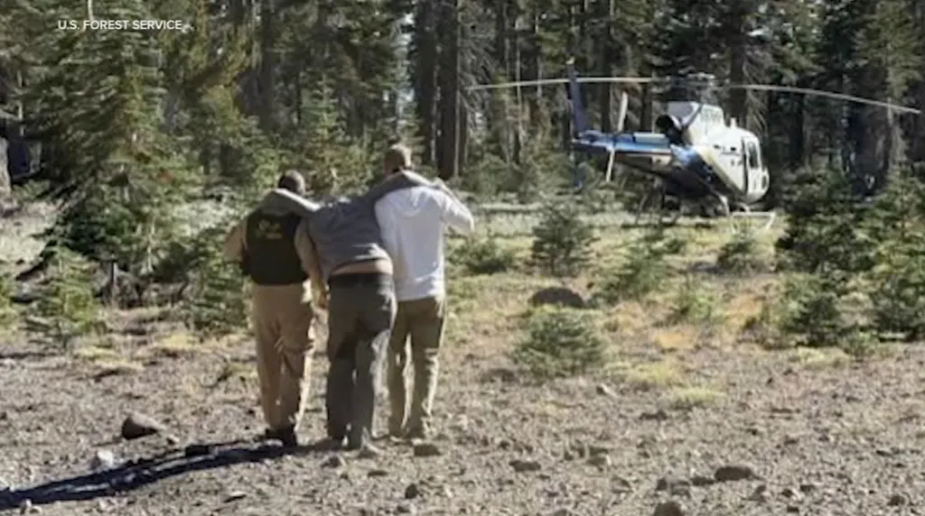 Two rescuers assist an injured climber toward a helicopter in a forest clearing.