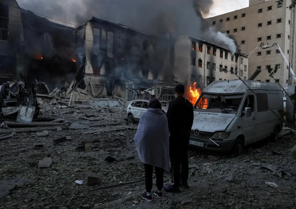 Residents stand at the site of a Russian missile attack in Kyiv, with destroyed buildings and vehicles, and fires burning.