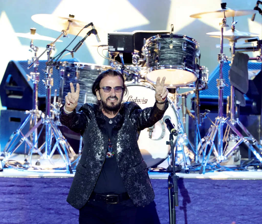 Ringo Starr on stage, wearing sunglasses, a sparkly black jacket, and a peace sign necklace, holding both hands up in a peace sign gesture.