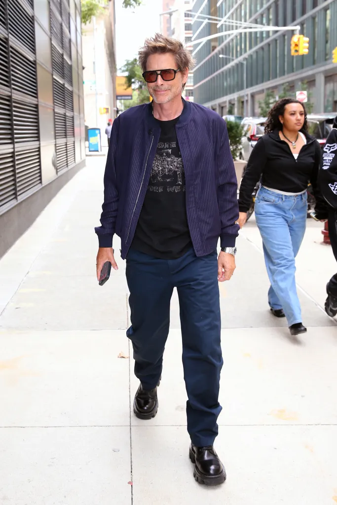 Rob Lowe walking on a city sidewalk in a navy blue bomber jacket, black t-shirt, and dark pants.
