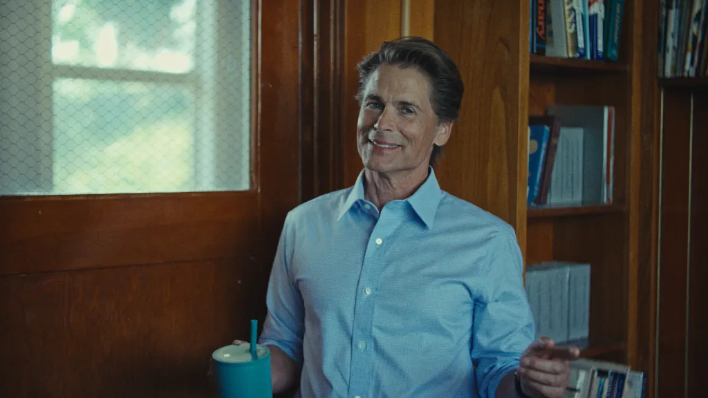 Rob Lowe in a light blue button-down shirt holding a teal cup.