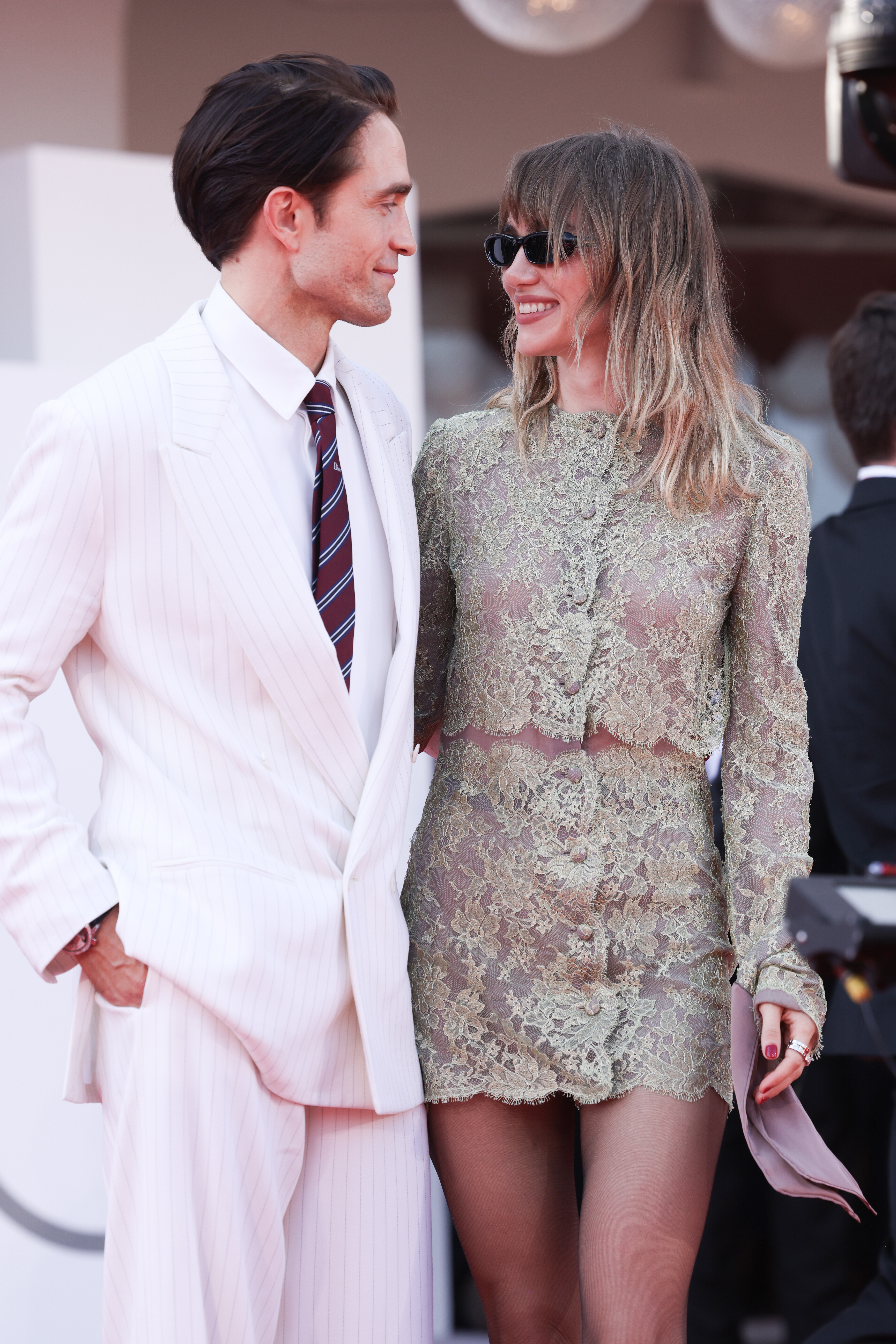 Robert Pattinson in an off-white pinstriped suit and Suki Waterhouse in a sheer, long-sleeved lace dress.