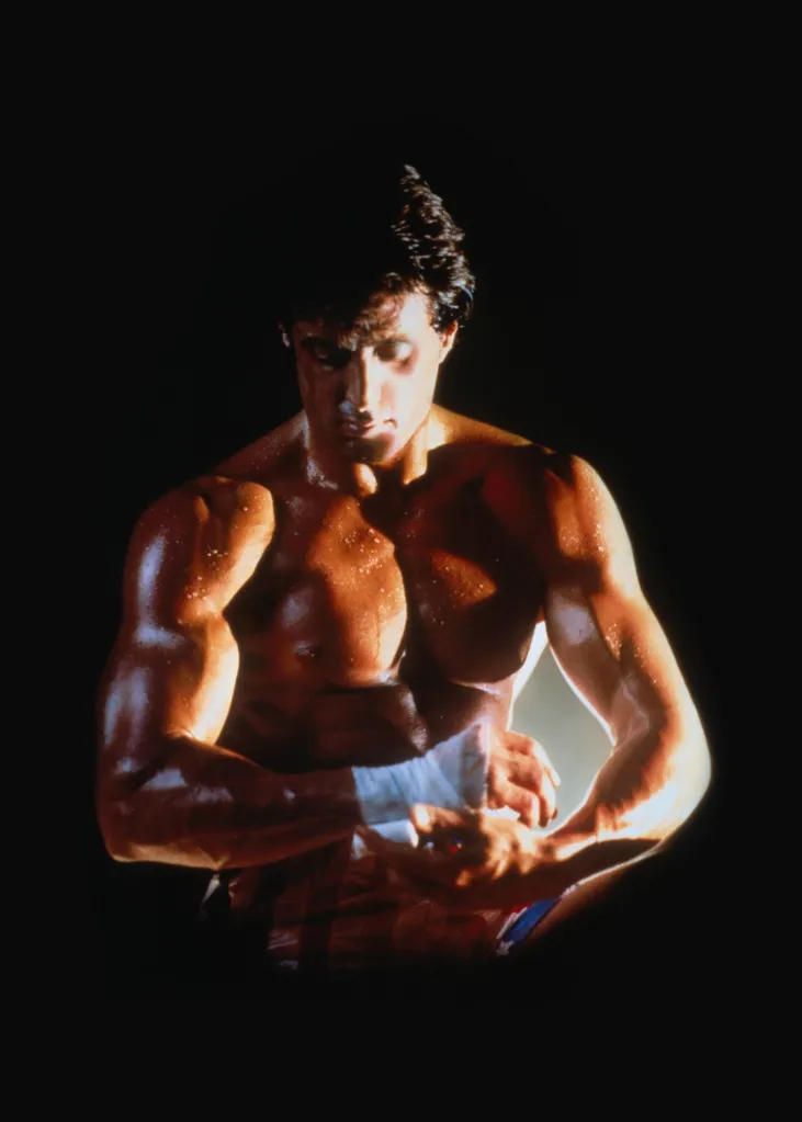 Sylvester Stallone as Rocky Balboa, barechested with dramatic lighting, wrapping his hands with white tape.