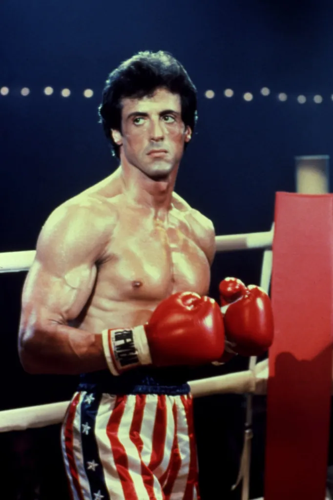 Sylvester Stallone as Rocky Balboa in a boxing ring, wearing red, white, and blue American flag boxing trunks and red gloves.