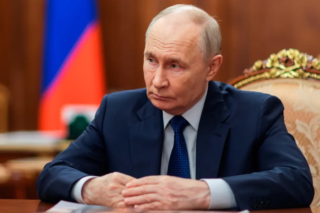 Russian President Vladimir Putin listens intently, wearing a dark suit and blue tie, with a Russian flag visible in the blurred background.