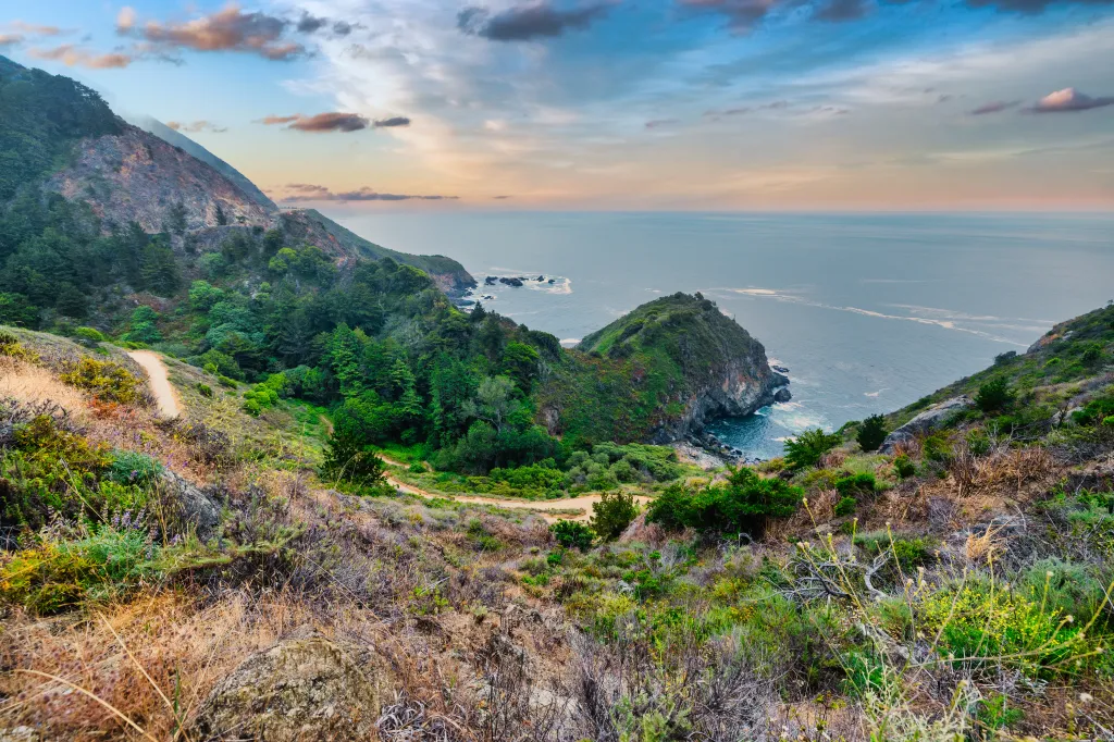 Scenic view of Partington Cove in Big Sur, California, featuring rugged cliffs, lush greenery, and the Pacific Ocean under a cloudy sky.
