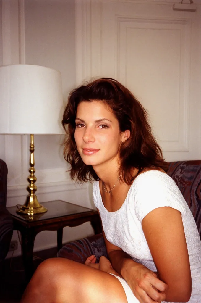 Sandra Bullock in a white dress, sitting in a chair next to a lamp.