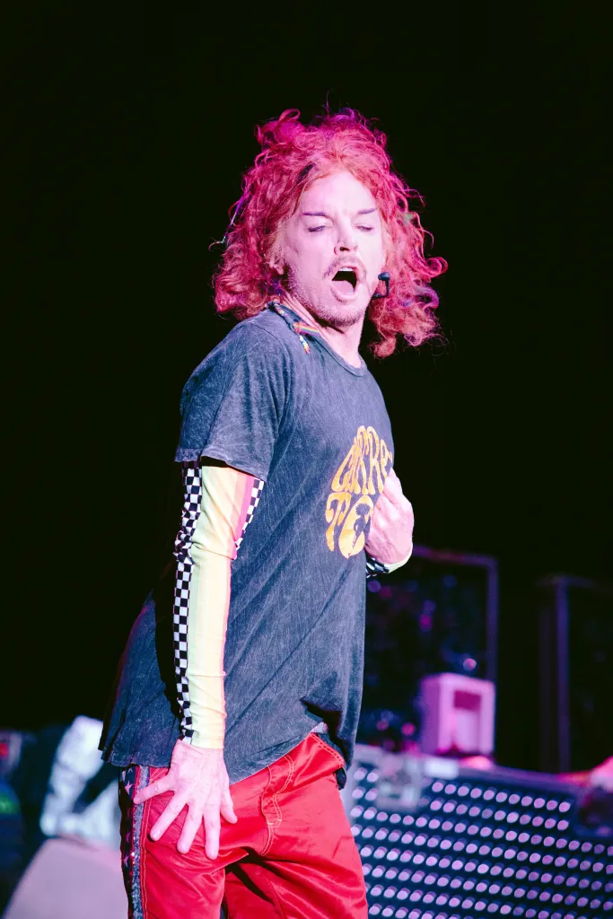 Comedian Carrot Top performing on stage in a black t-shirt, red pants, and colorful sleeves.