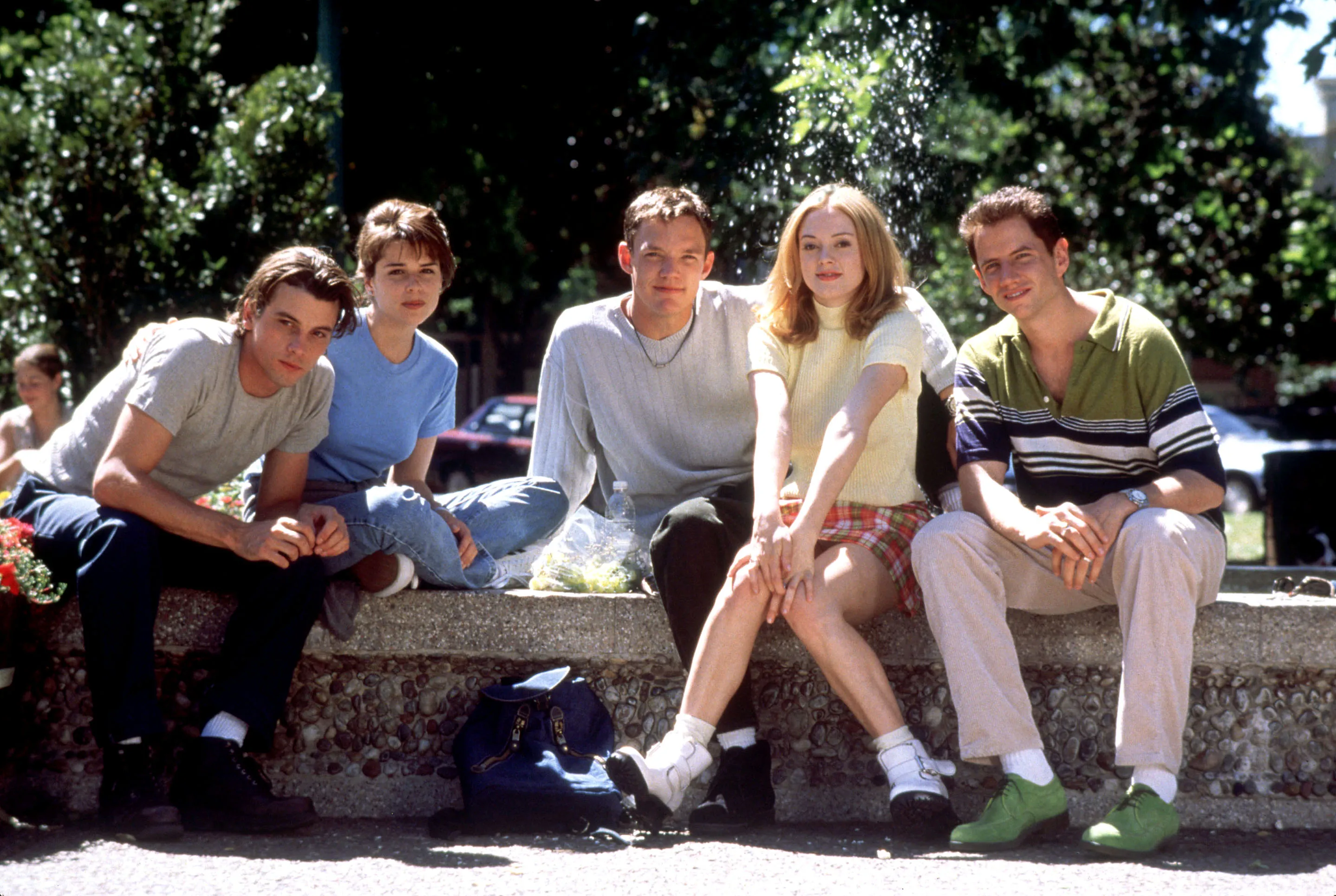 Skeet Ulrich, Neve Campbell, Matthew Lillard, Rose McGowan, and Jamie Kennedy sitting together on a low wall outdoors.