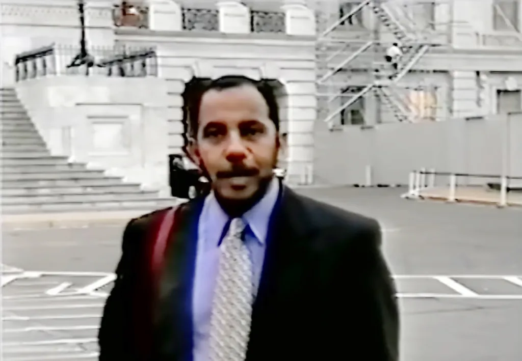 A man with a mustache and dark hair, wearing a suit and light blue shirt with a patterned tie, stands outdoors in front of a white building with stairs.