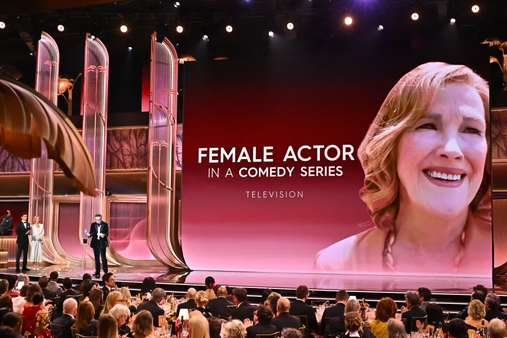 Seth Rogen presenting at an awards ceremony, with Catherine O'Hara visible on the screen behind him, nominated for 