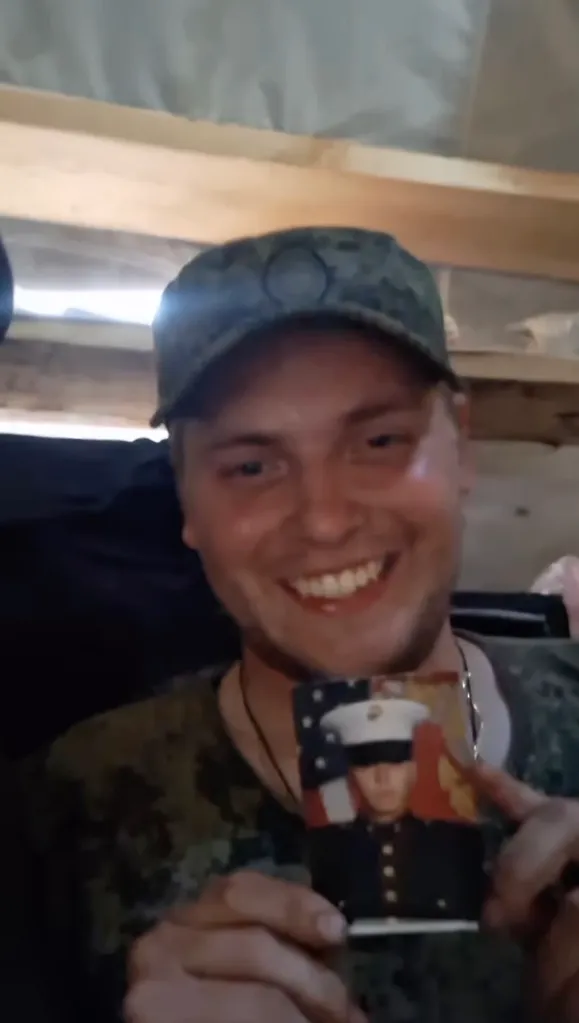 Seth William Baker, smiling, holding up a picture of himself in a US Marine uniform with an American flag background.