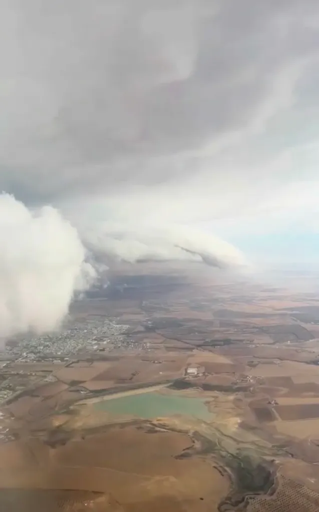 View from an airplane window showing storm clouds gathering over a rural landscape with a small town and a light green lake.