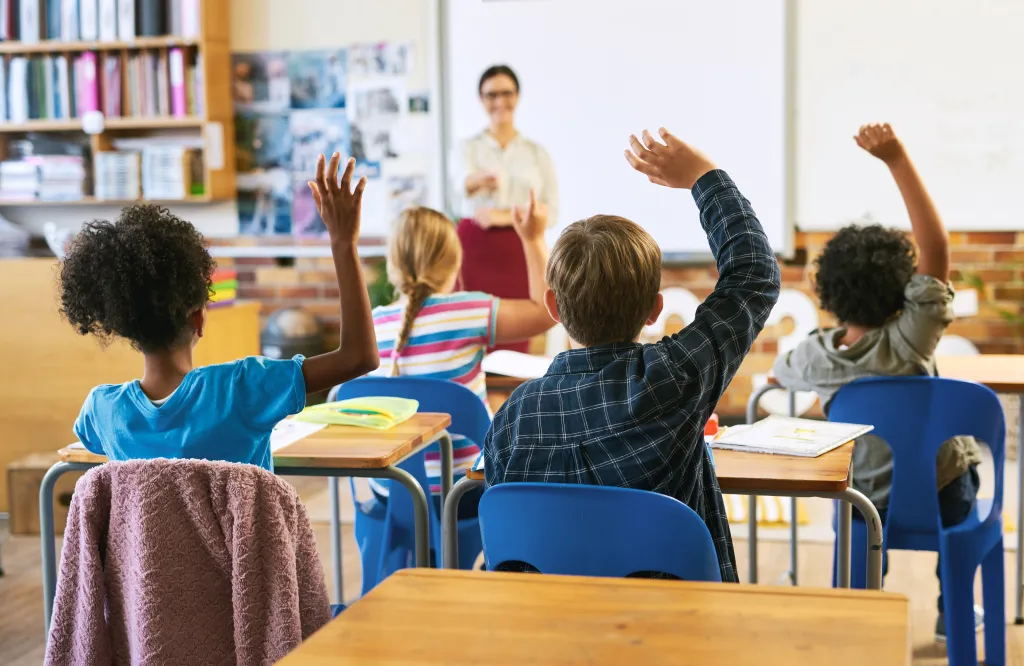 Children raising their hands in a classroom, with a teacher in the background.