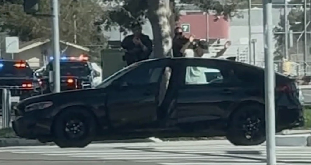 A black car with open doors and several police cars with flashing lights in the background.