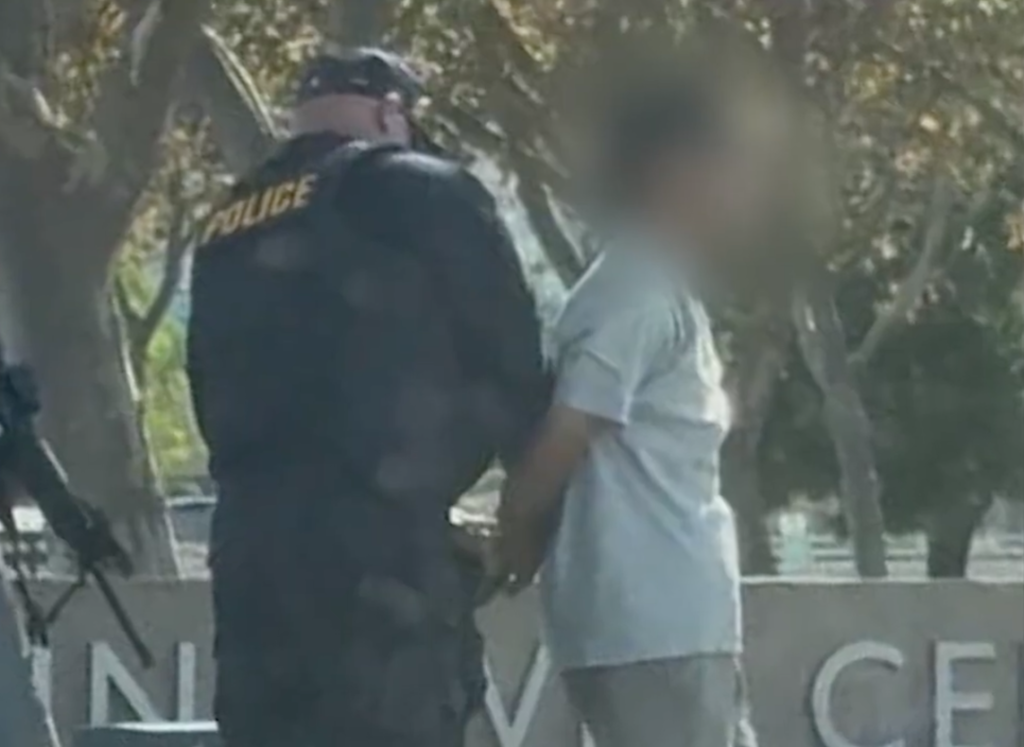 A police officer facing away from the camera, standing next to a blurred figure with their hands handcuffed behind their back.