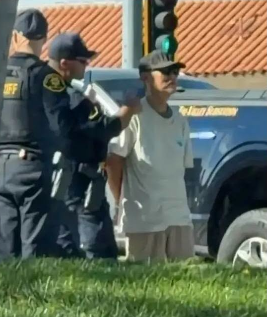 Police officers detain a man in handcuffs by a vehicle in a parking lot.