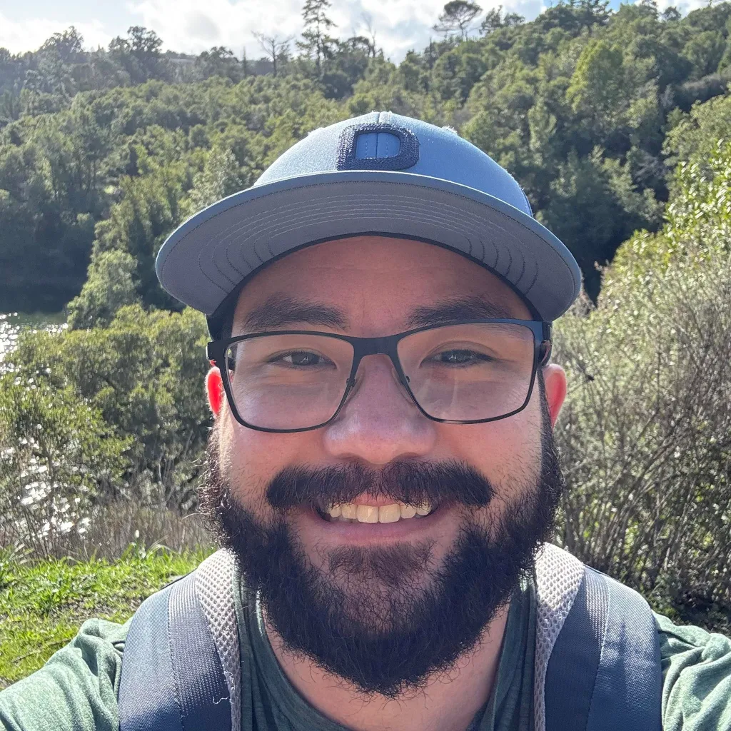 Jonathan McKinsey wearing a blue baseball cap, glasses, and a beard, smiling at the camera outdoors.